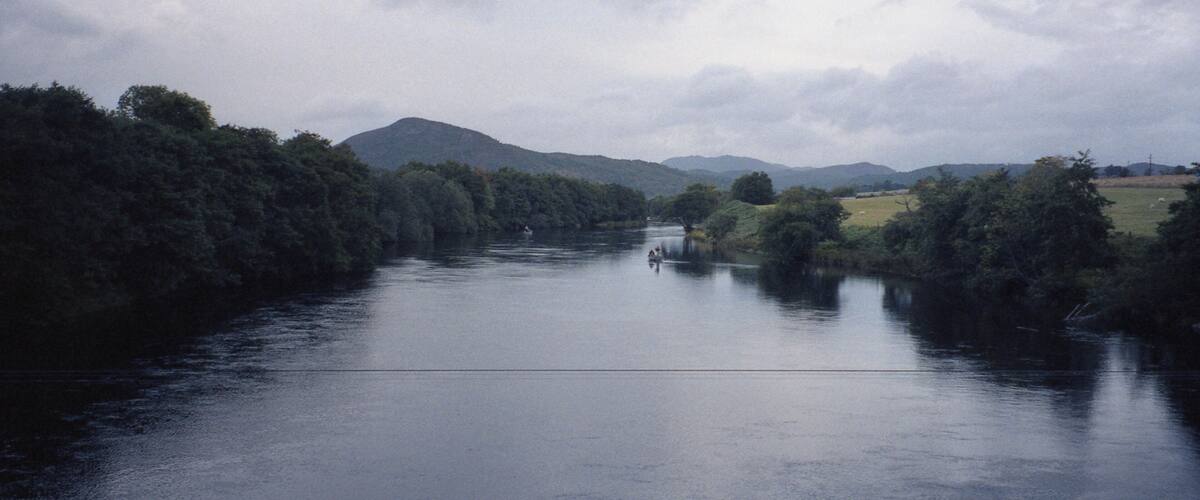 A river flanked by dense forests and distant mountains under a cloudy sky.