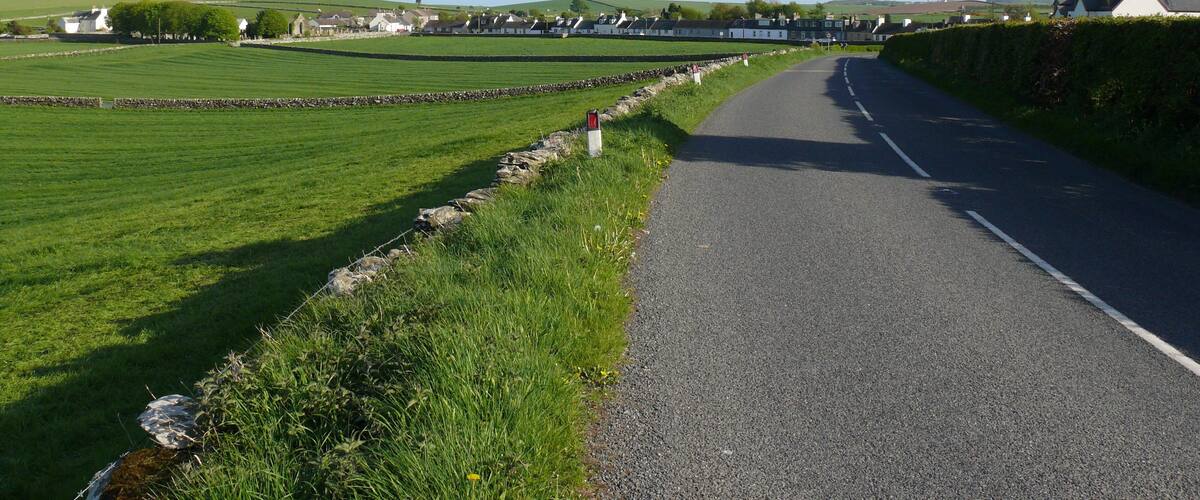 A rural road flanked by stone walls and fields, leading to a cluster of houses.