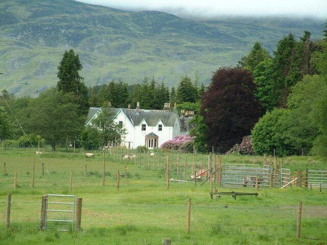 A white house with a green roof, surrounded by a fenced field with sheep, set against a mountainous backdrop.
