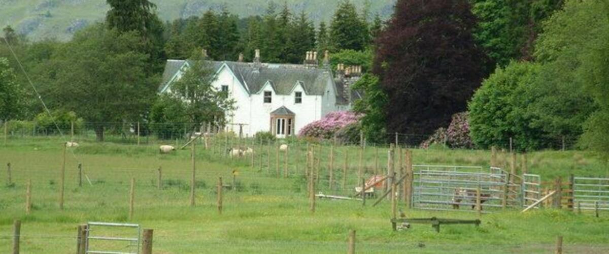 A white house with a green roof, surrounded by a fenced field with sheep, set against a mountainous backdrop.