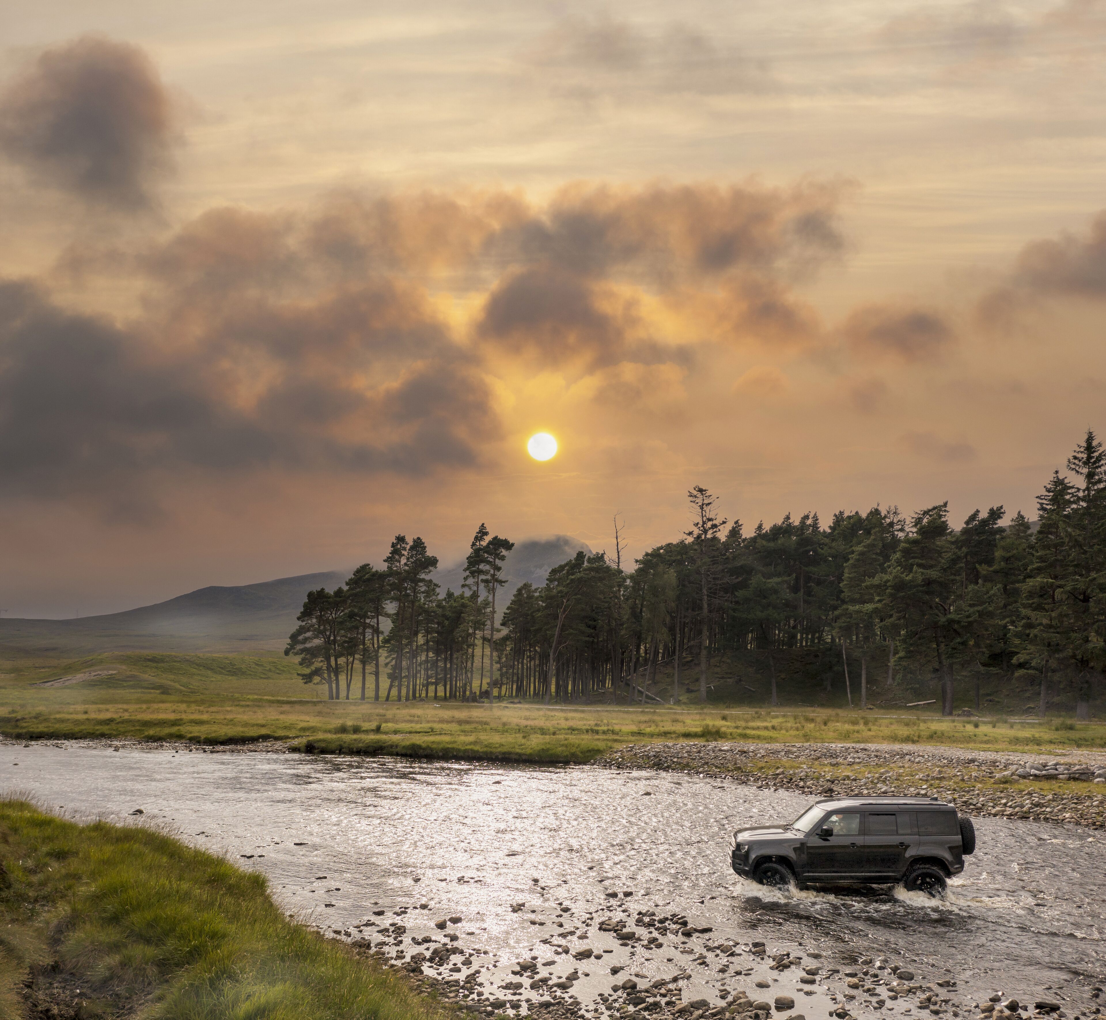 Aerial view of a vehicle forging the river at Ardverikie Estate amidst the tranquil Scottish Highlands at sunset, Newtonmore, Scotland, United Kingdom.