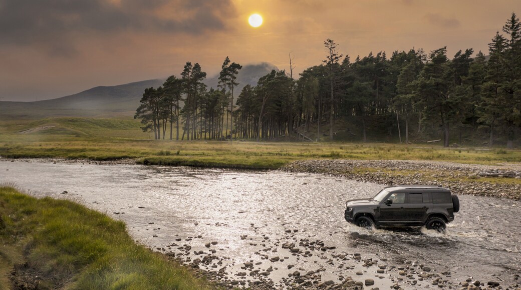 Aerial view of a vehicle forging the river at Ardverikie Estate amidst the tranquil Scottish Highlands at sunset, Newtonmore, Scotland, United Kingdom.