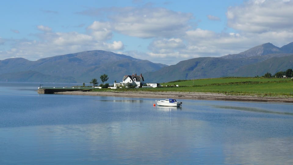 A calm lake with a small boat, a white house, and mountains in the background.