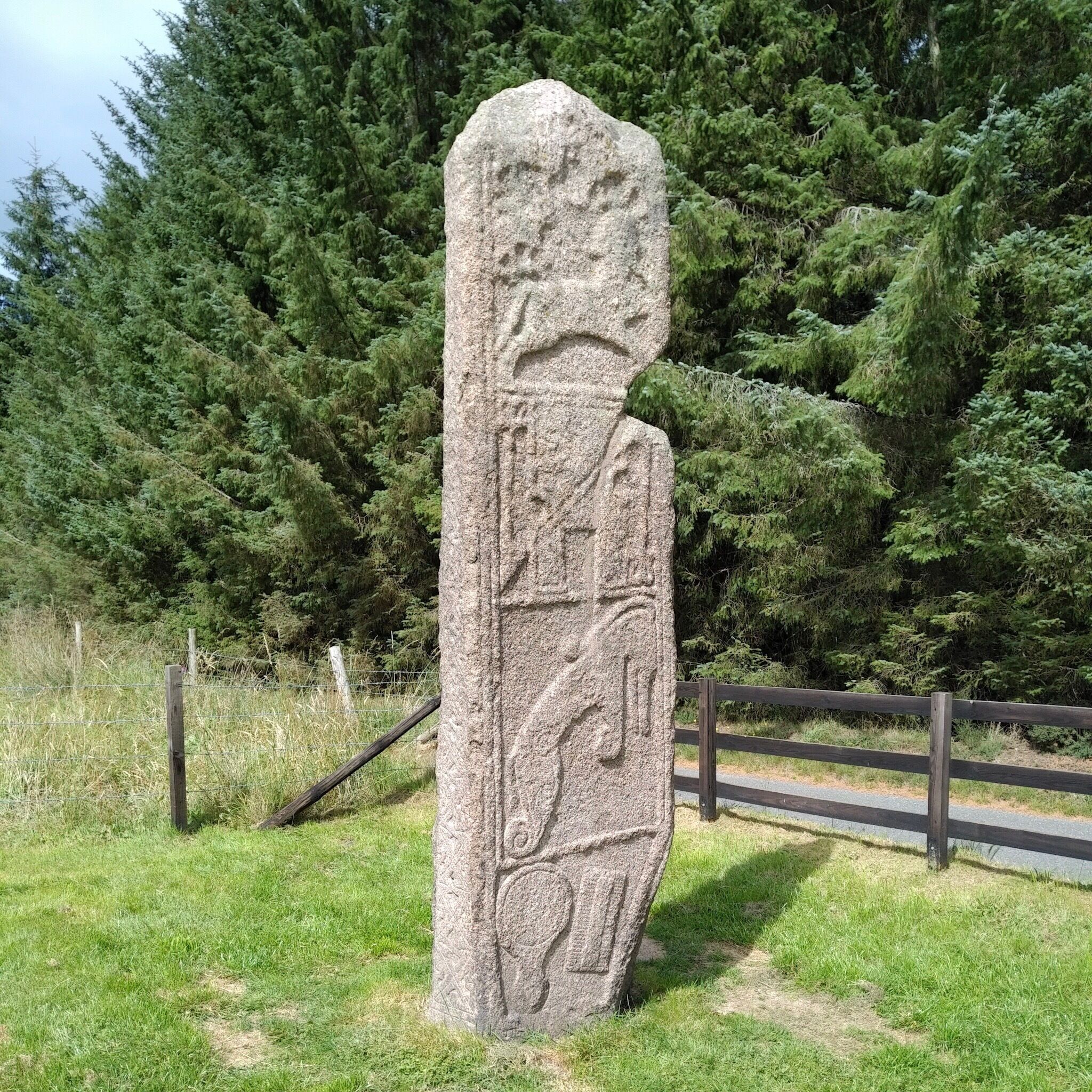 A large stone monument with carvings, surrounded by grass and a wooden fence, with dense forest in the background.
