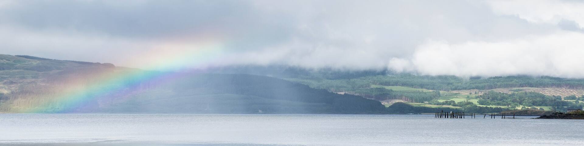 A lake with a rainbow, forested hills, and a cloudy sky.