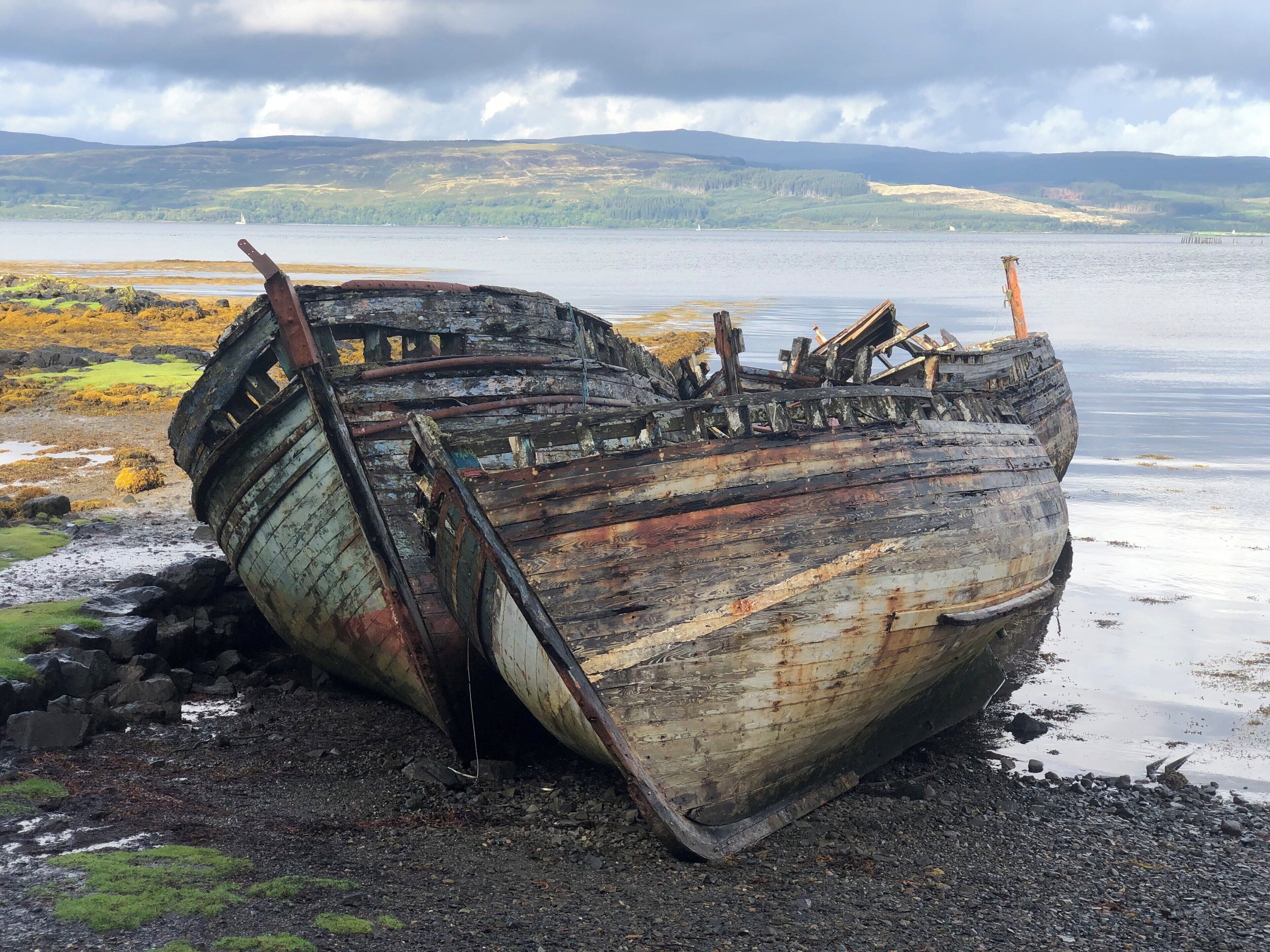 A rusted, abandoned shipwreck on a rocky shore with a lake and distant hills in the background.
