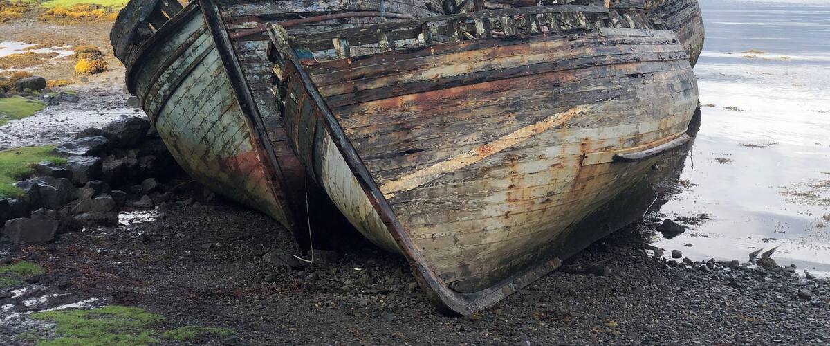 A rusted, abandoned shipwreck on a rocky shore with a lake and distant hills in the background.