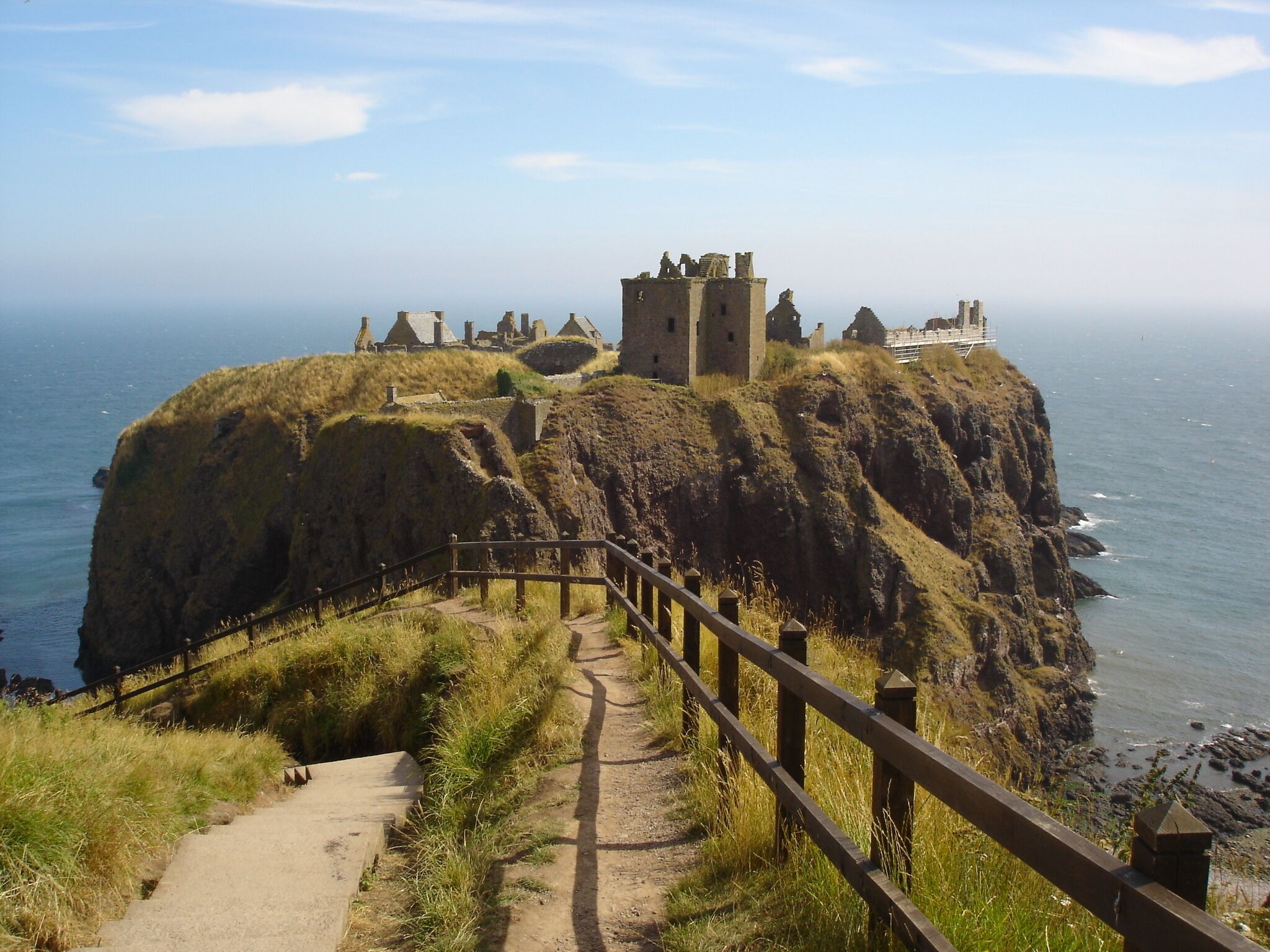 Um penhasco costeiro com um castelo histórico e ruínas, uma passarela de madeira e o oceano.