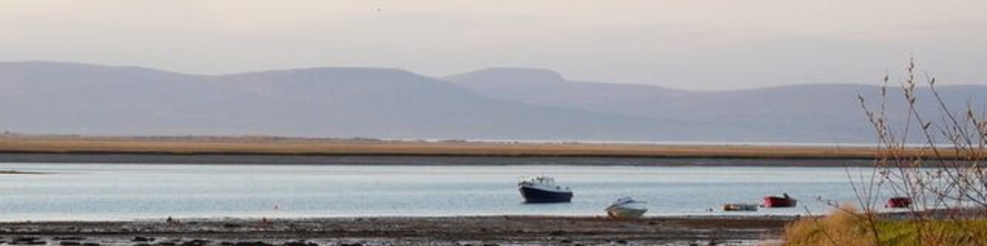Paisagem costeira com um barco na água, uma praia de areia e montanhas ao longe.