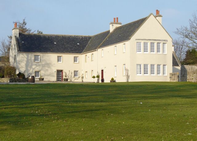 A large, two-story house with a sloped roof and multiple chimneys, surrounded by a well-maintained lawn.