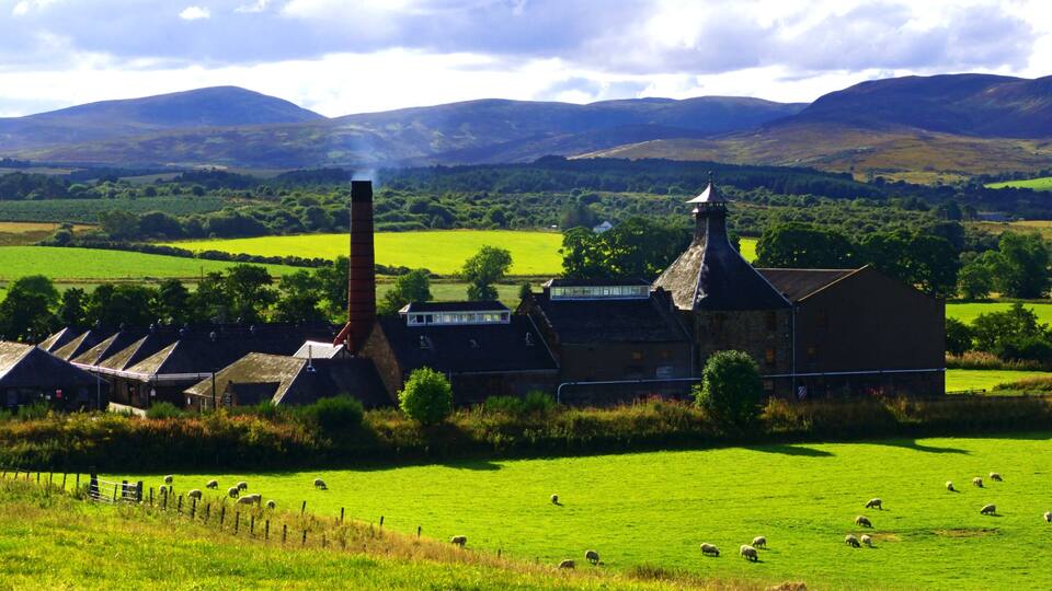 A rural landscape with a stone building, sheep grazing, and rolling hills in the background.