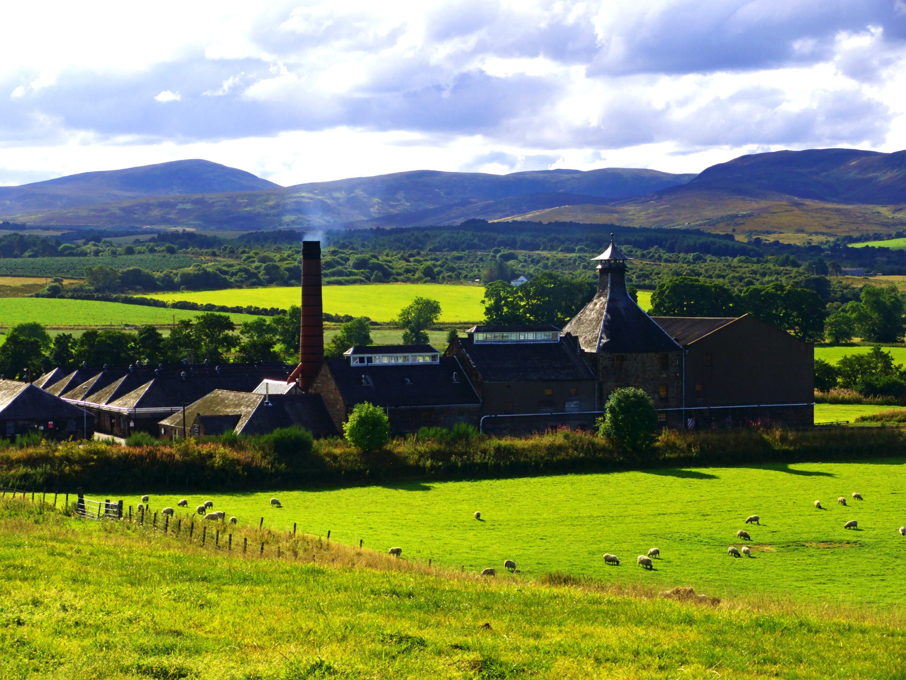 A rural landscape with a stone building, sheep grazing, and rolling hills in the background.