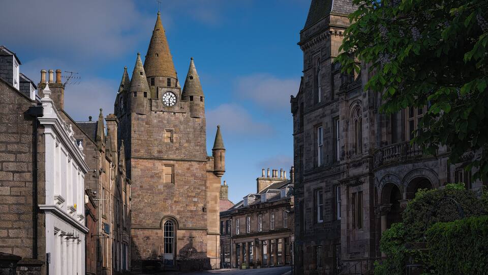 A historic stone tower with a clock, surrounded by traditional buildings and a clear blue sky.