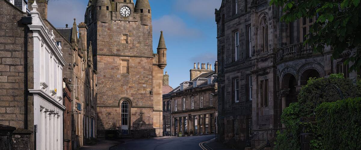 A historic stone tower with a clock, surrounded by traditional buildings and a clear blue sky.