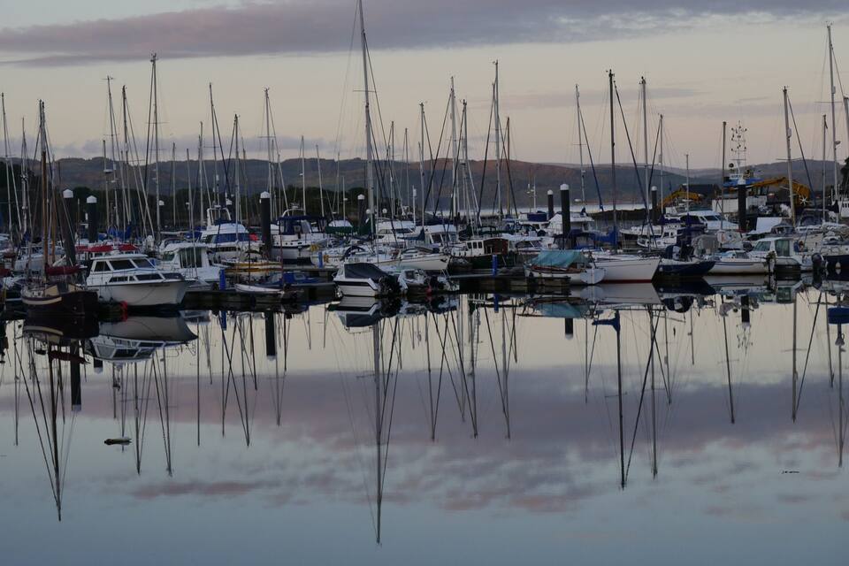A marina with numerous sailboats docked, surrounded by calm water and distant hills.