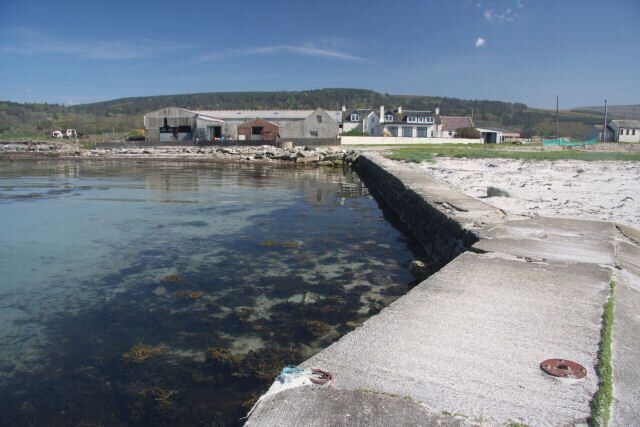A coastal area with a concrete pier extending into a clear water body, surrounded by buildings and hills in the background.