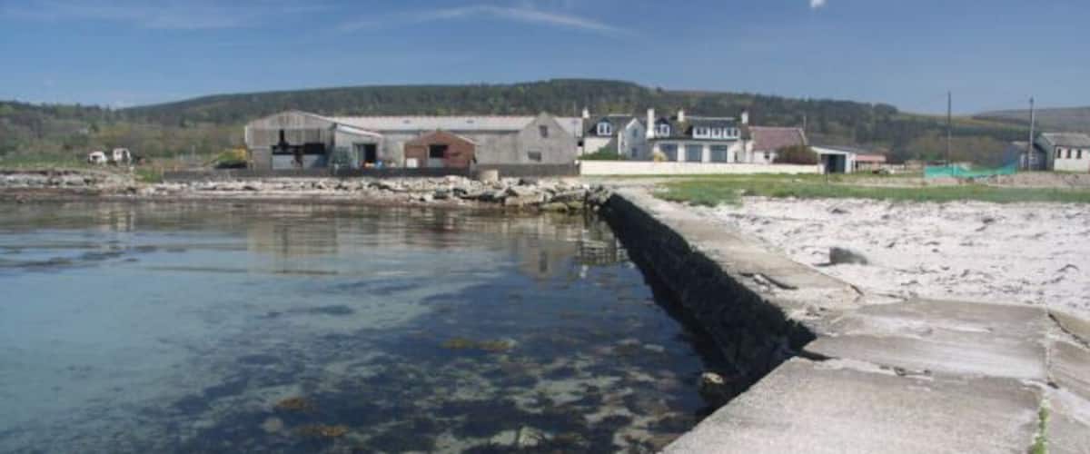 A coastal area with a concrete pier extending into a clear water body, surrounded by buildings and hills in the background.