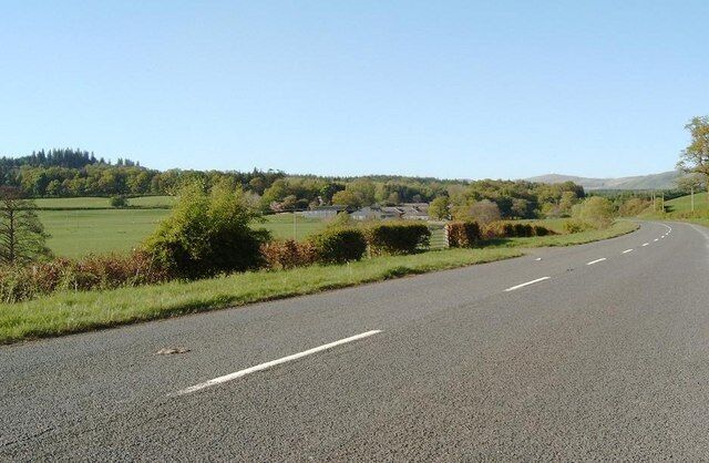 A rural road with a clear sky, green fields, and distant buildings.