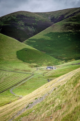 A rural landscape with rolling hills, a winding road, and a small house.