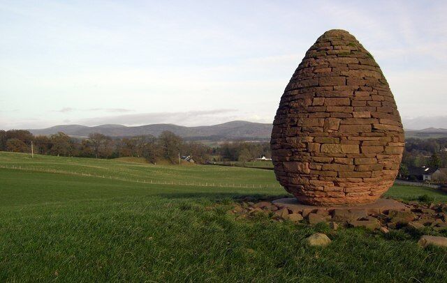 A large, egg-shaped stone structure in a grassy field with a backdrop of hills and a clear sky.