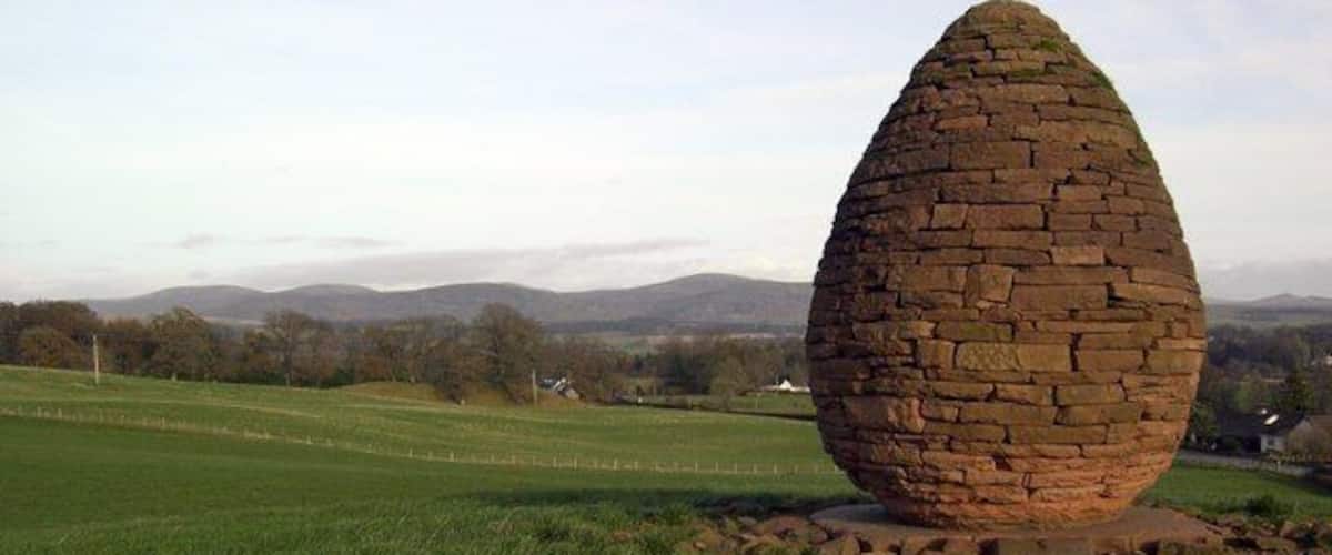 A large, egg-shaped stone structure in a grassy field with a backdrop of hills and a clear sky.