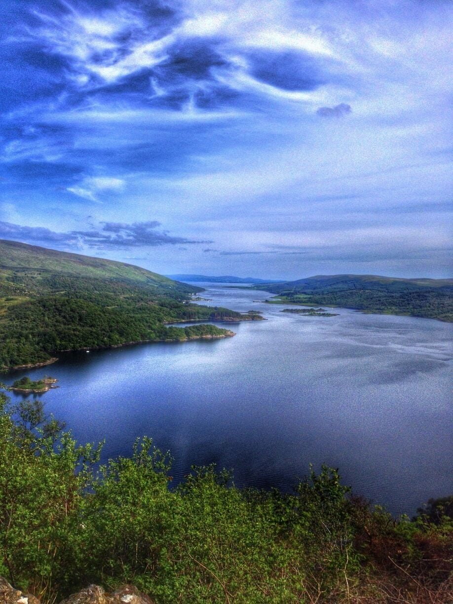 A large body of water surrounded by green hills and a cloudy sky.