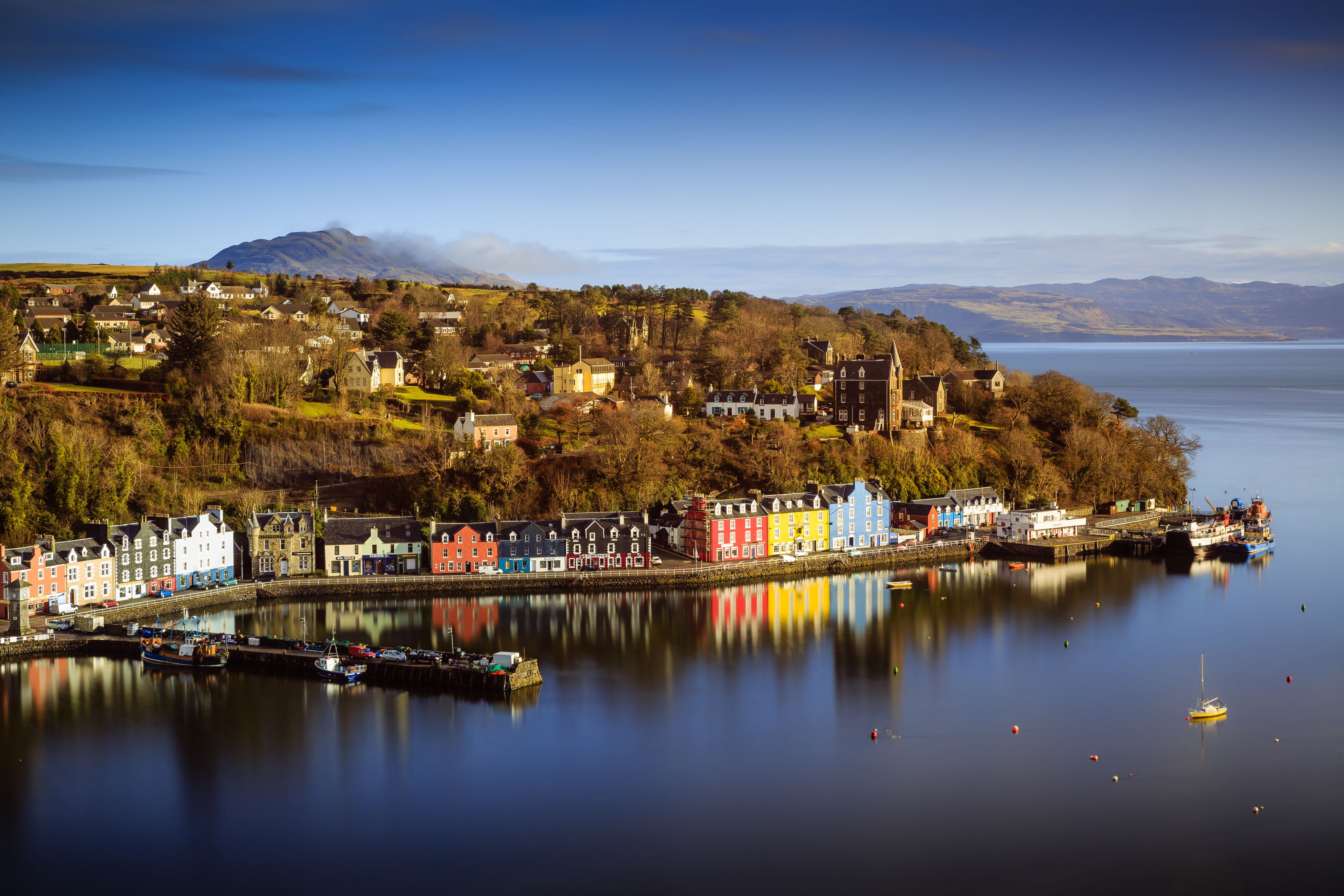 A coastal town with colorful buildings, a calm harbor, and a mountain in the background.