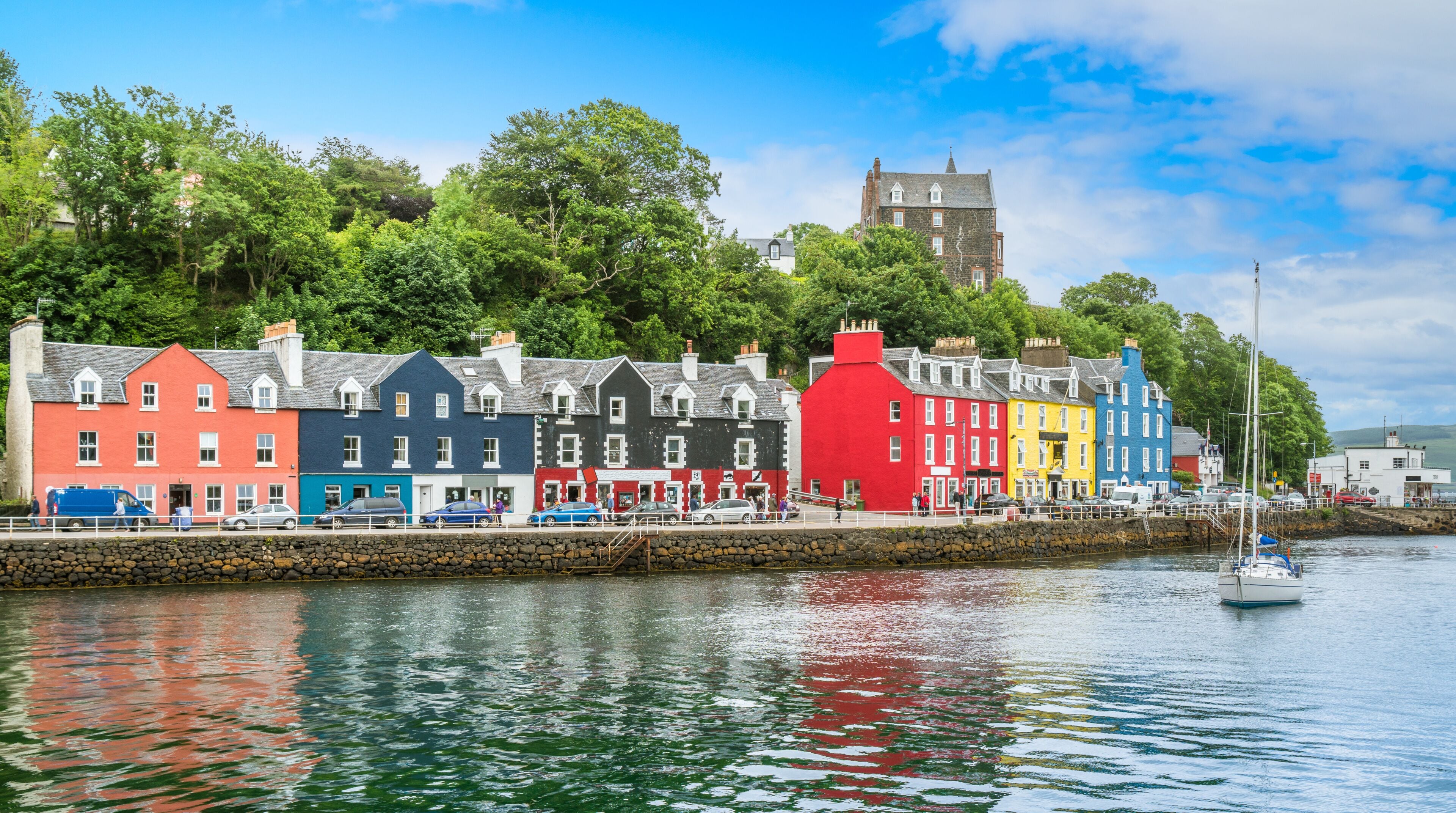 A waterfront with colorful buildings, a stone wall, and a sailboat.