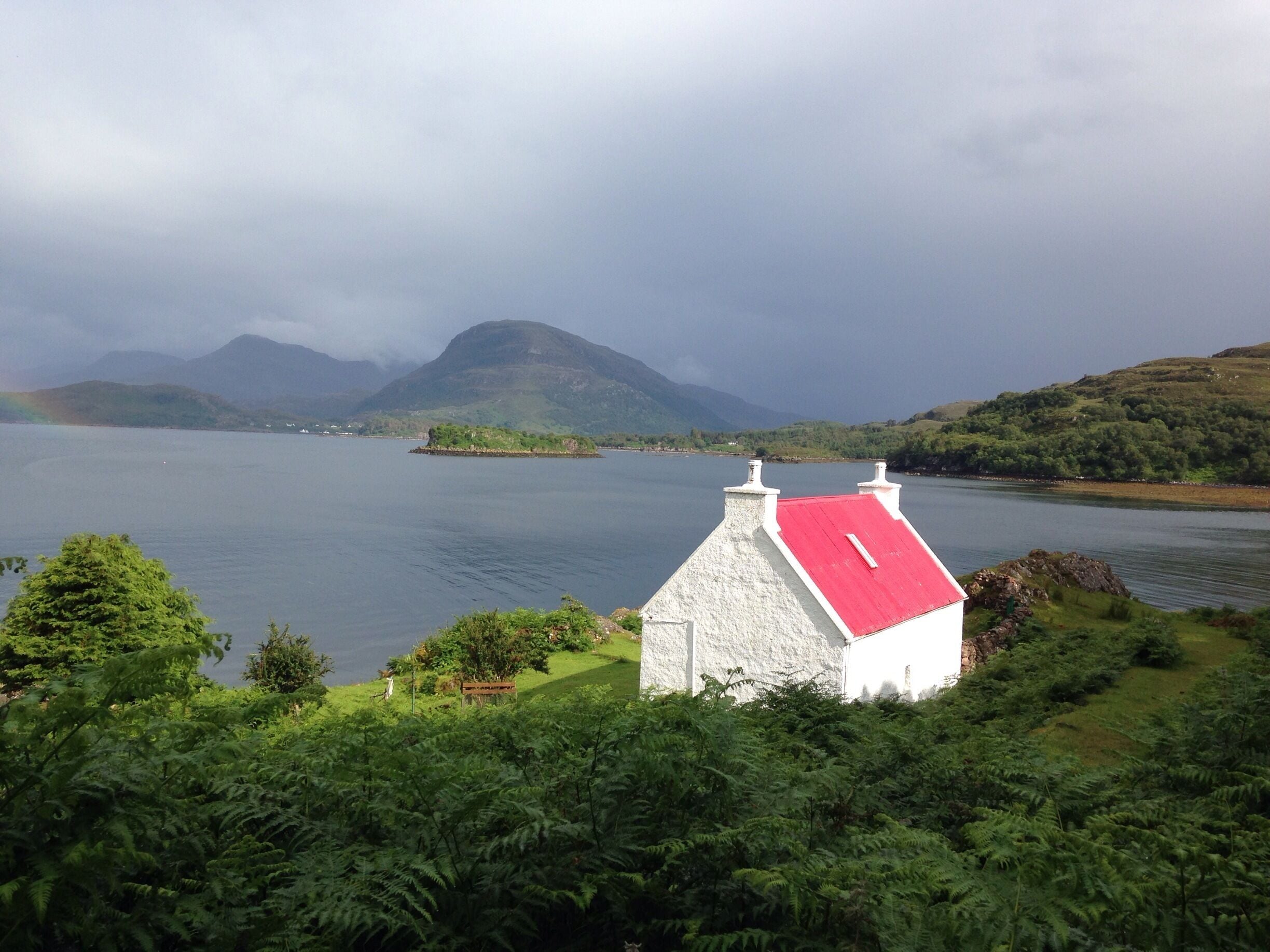 A white house with a red roof, a lake, and mountains in the background.
