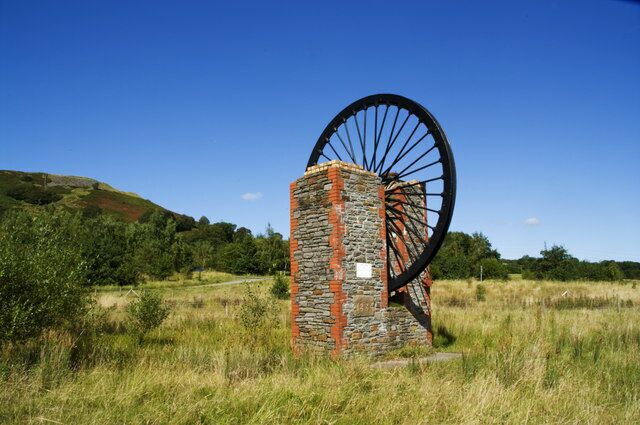 A windmill with a stone base and large black wheel in a grassy field with hills in the background.