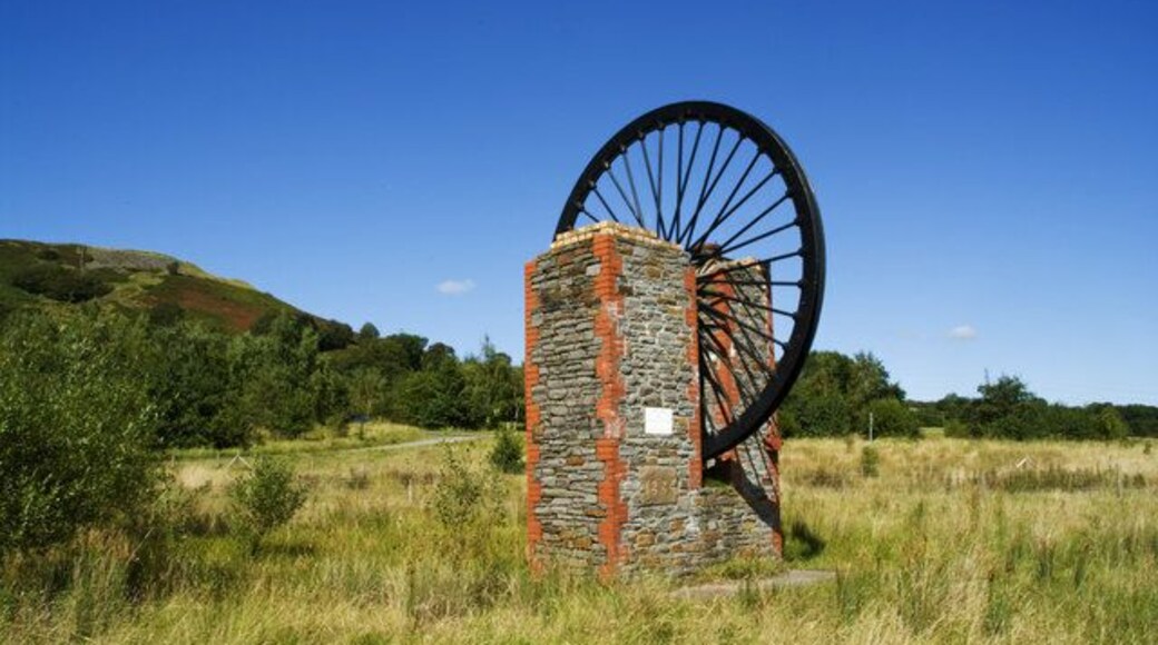 A windmill with a stone base and large black wheel in a grassy field with hills in the background.