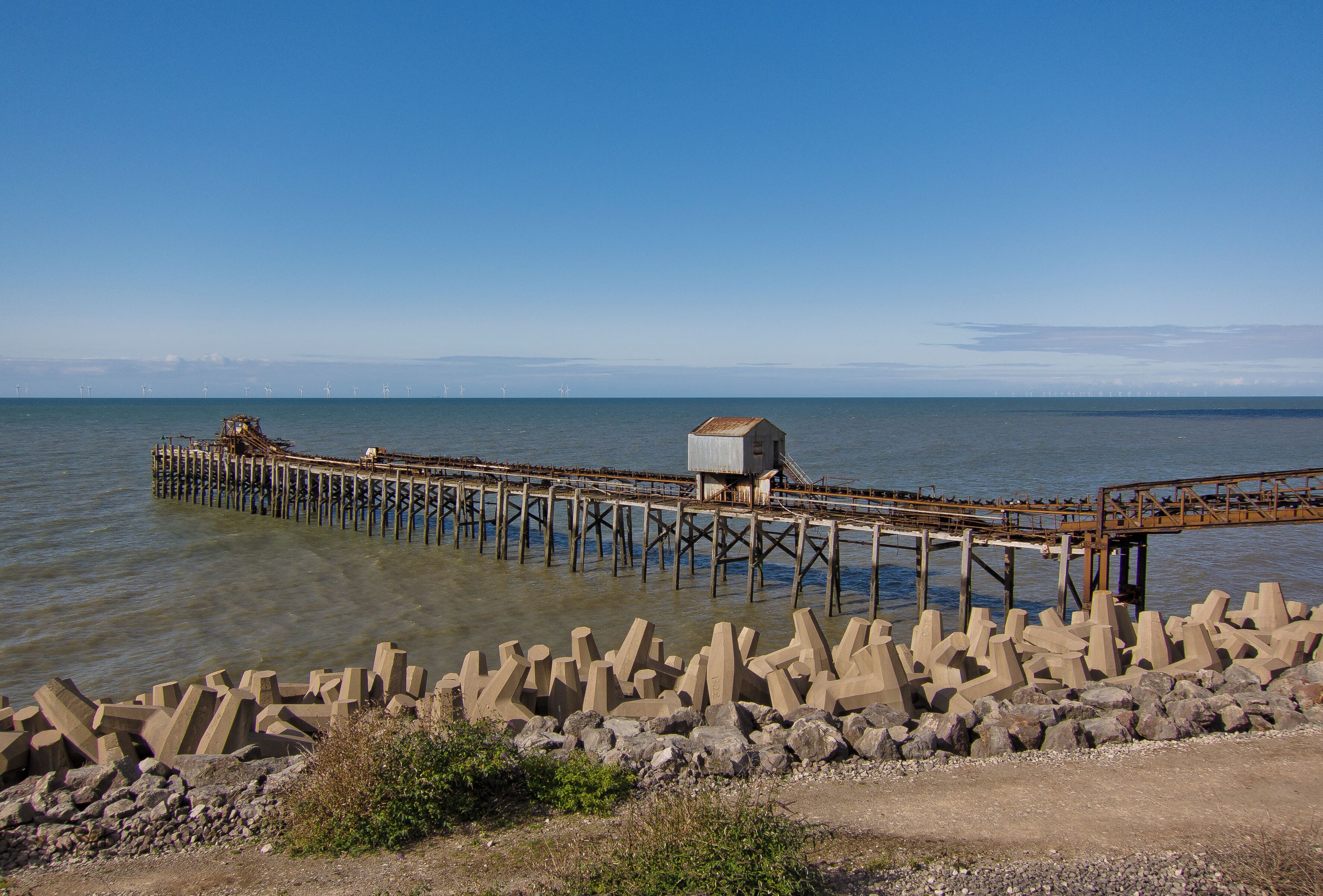 A pier extending into calm waters, a small building on the pier, and a rocky breakwater in the foreground.