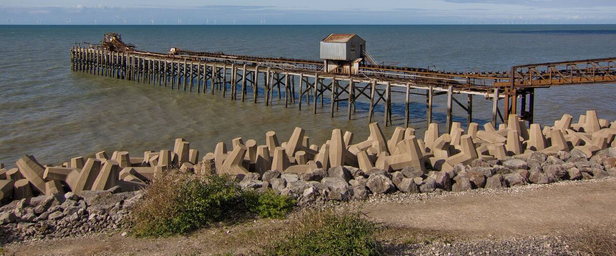 A pier extending into calm waters, a small building on the pier, and a rocky breakwater in the foreground.