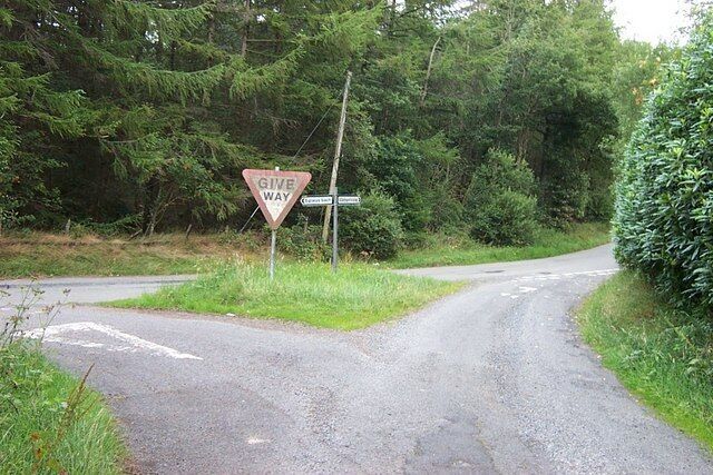 A rural road with a 'Give Way' sign at a junction, surrounded by dense greenery.