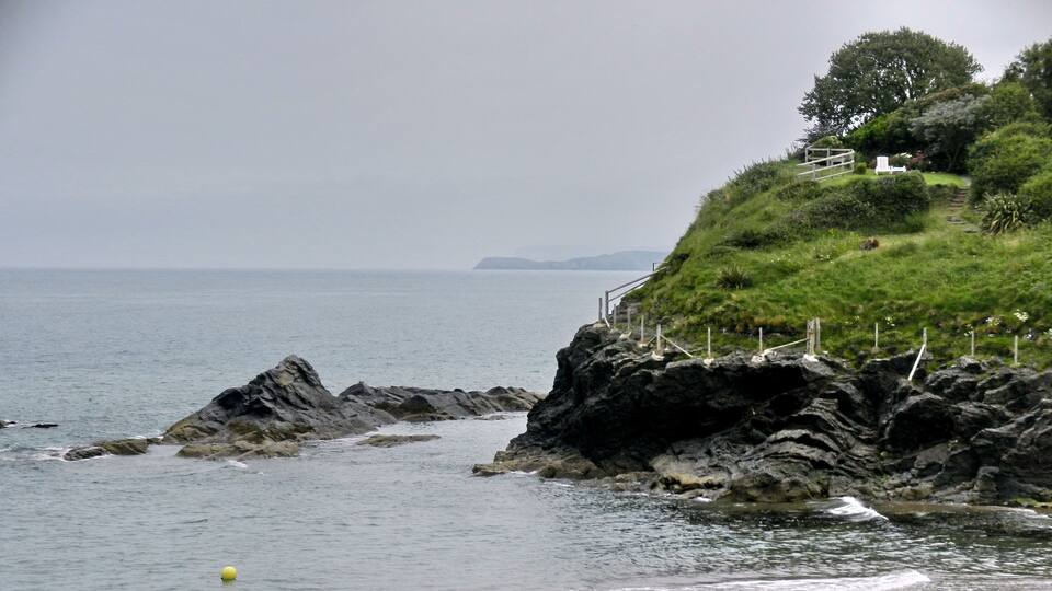 A rocky coastline with a sandy beach, a grassy hill with a fence, and a child playing near the water.