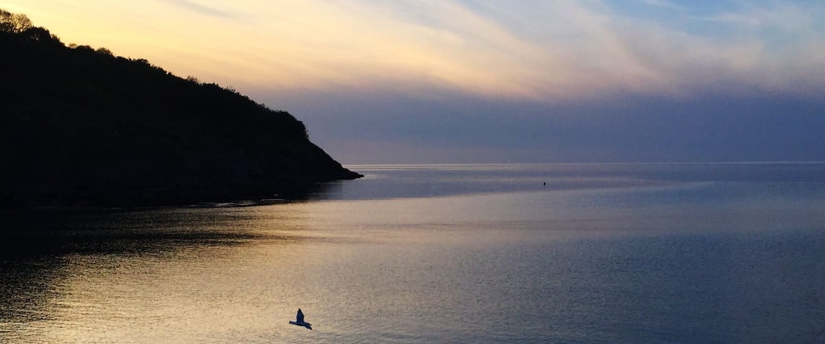 A calm sea at sunset with a beach and a hill in the background.