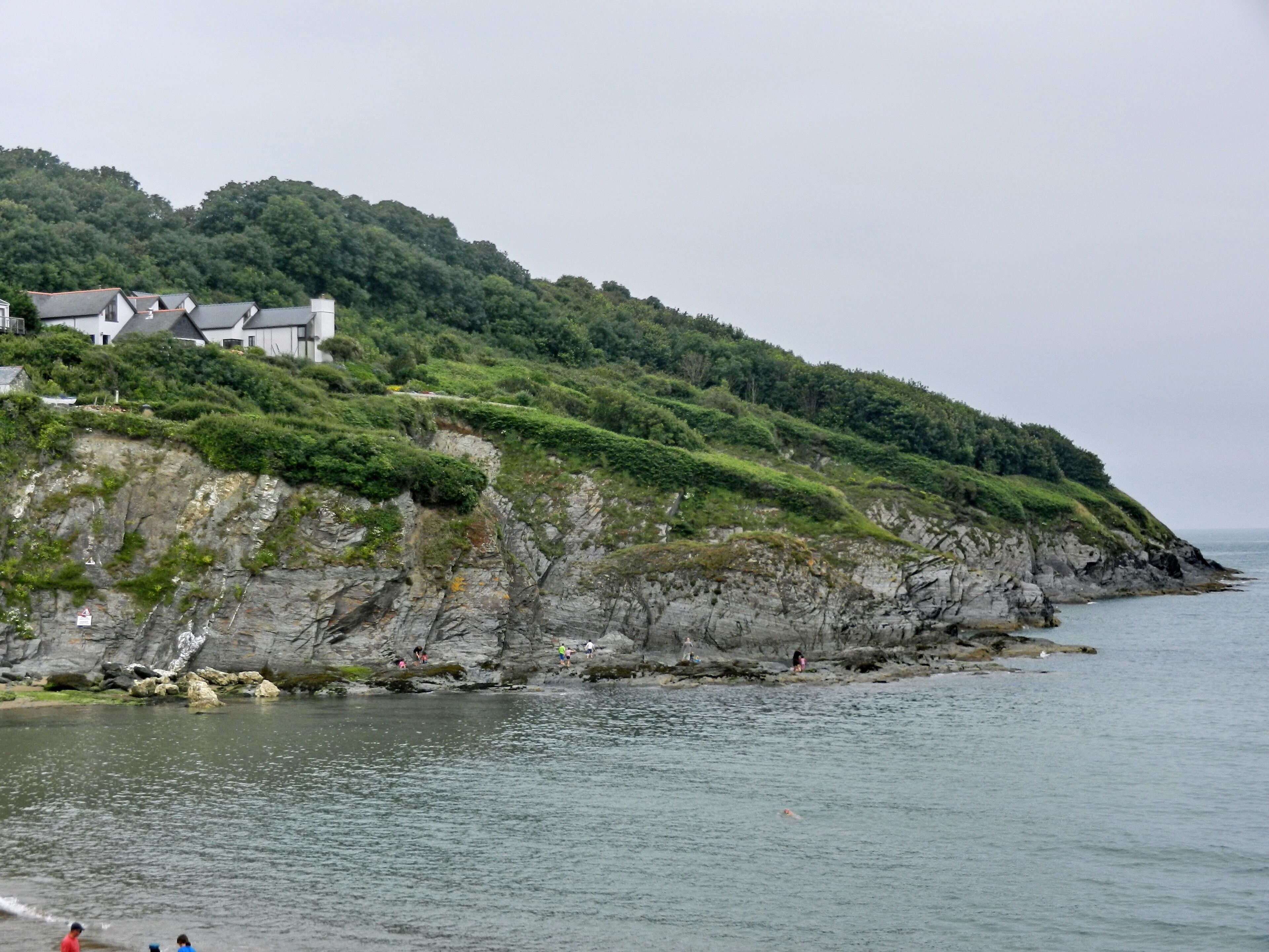 A coastal landscape with a rocky cliff, lush greenery, and a building on the hillside.