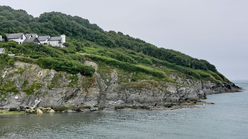 A coastal landscape with a rocky cliff, lush greenery, and a building on the hillside.