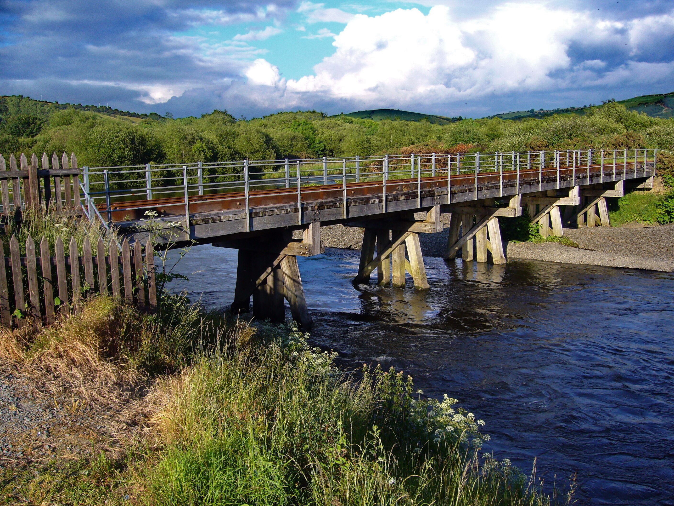A wooden bridge over a river with a metal railing, surrounded by greenery and hills.