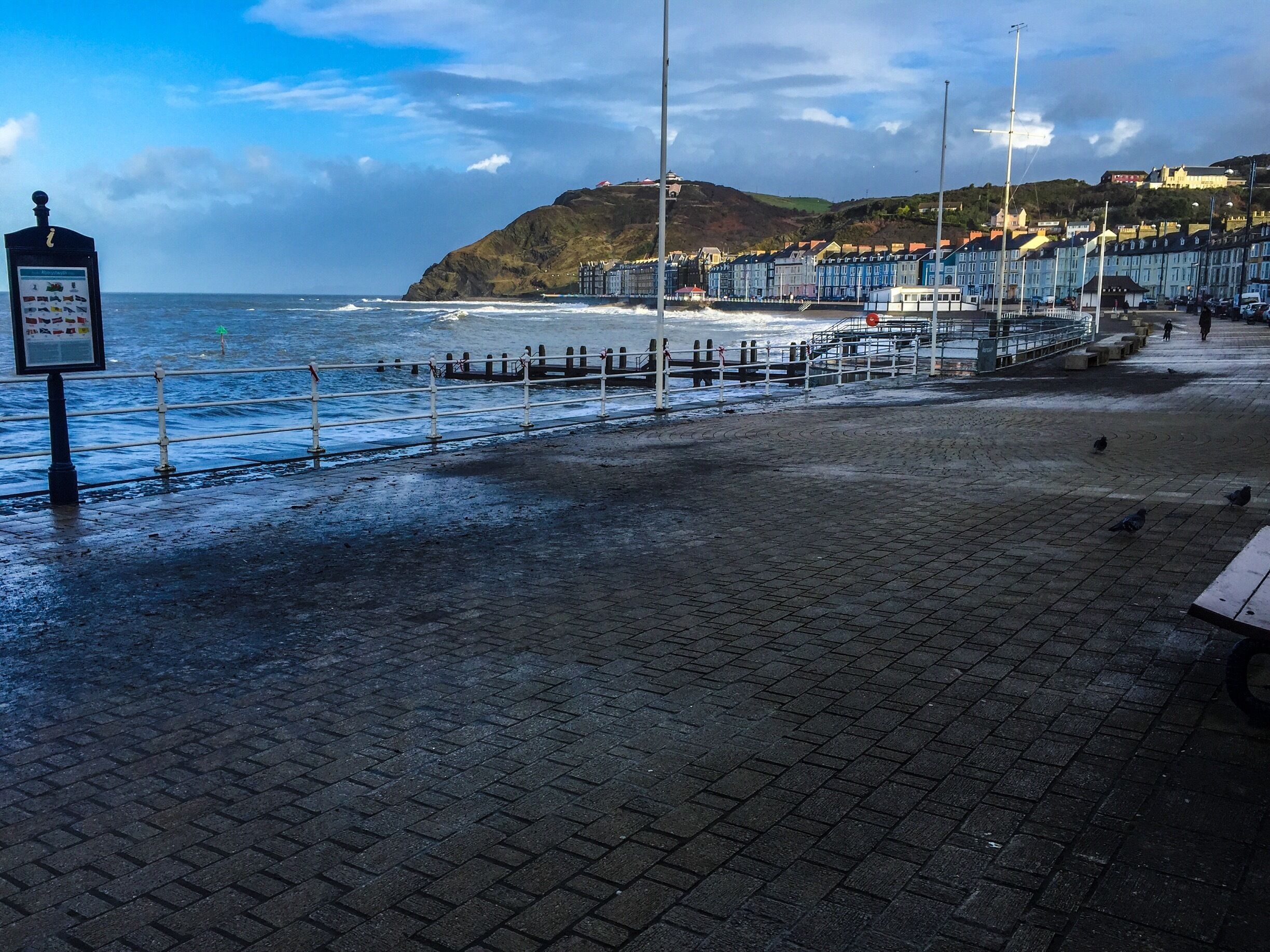 A coastal promenade with a sea wall, a pier, and buildings along the hillside.
