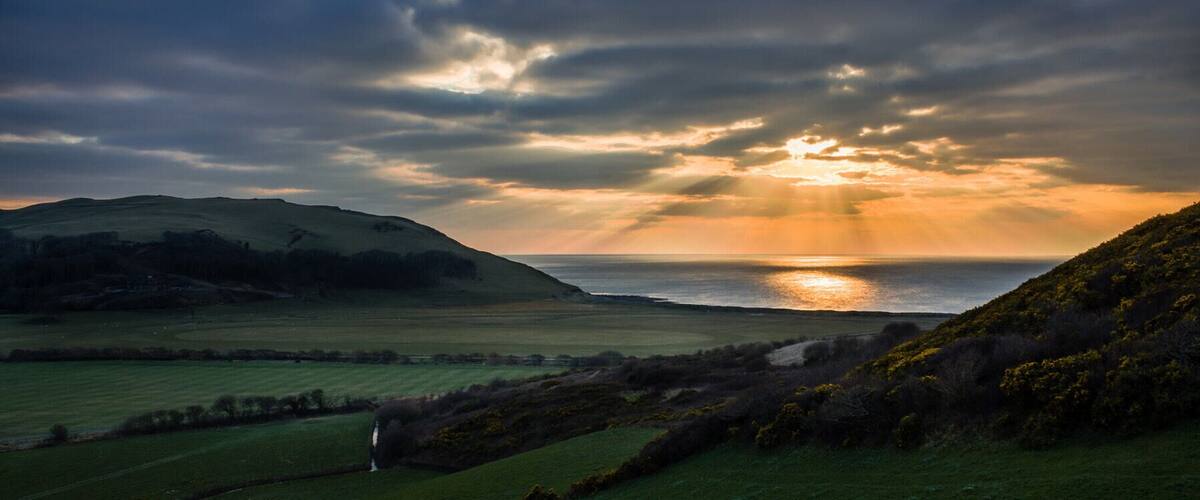A coastal landscape with rolling hills, a body of water, and a sunset.