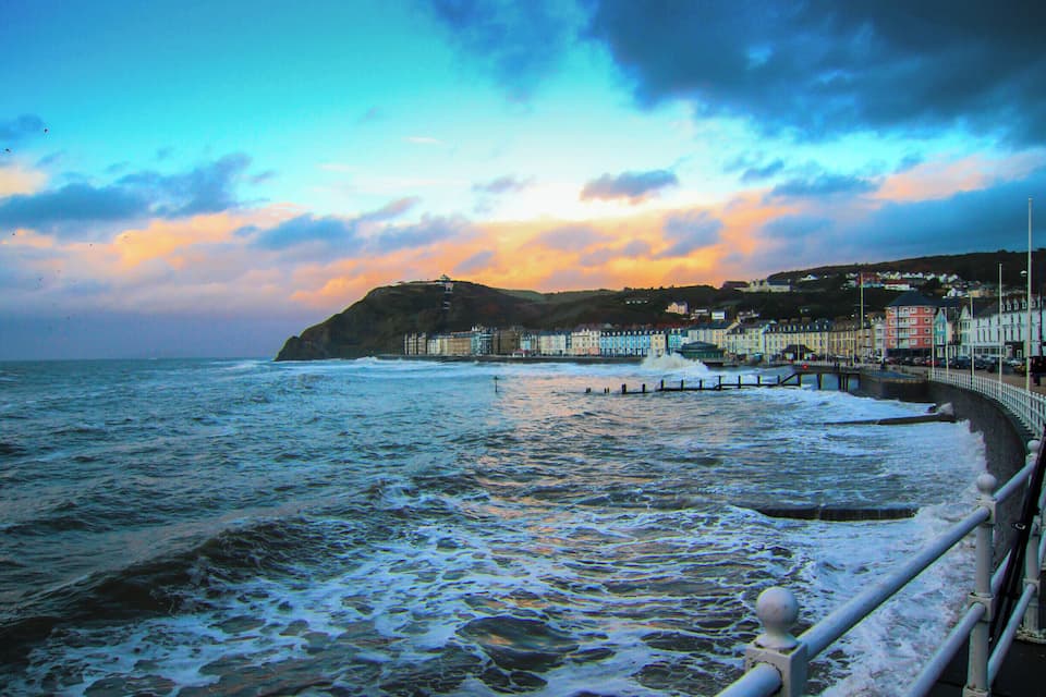 A coastal town with a promenade, waves crashing onto the shore, and a sunset sky.
