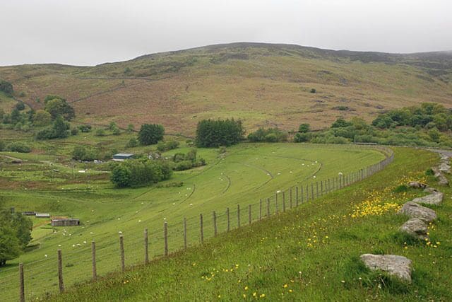 A green field with a fence, a small building, and rolling hills in the background.
