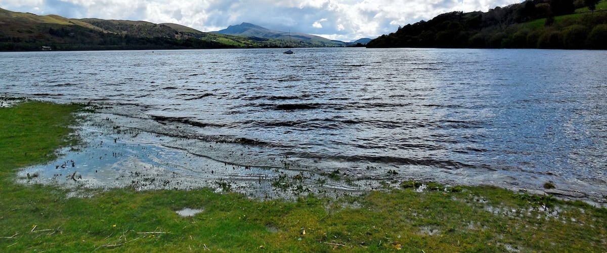 Un lac où navigue un voilier, entouré de collines et surplombé d’un ciel partiellement nuageux.