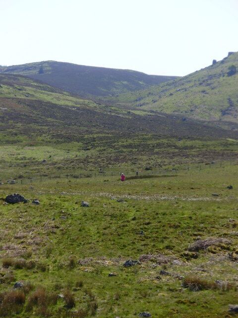Un campo cubierto de hierba con una persona vestida de rojo, una colina al fondo y rocas dispersas.
