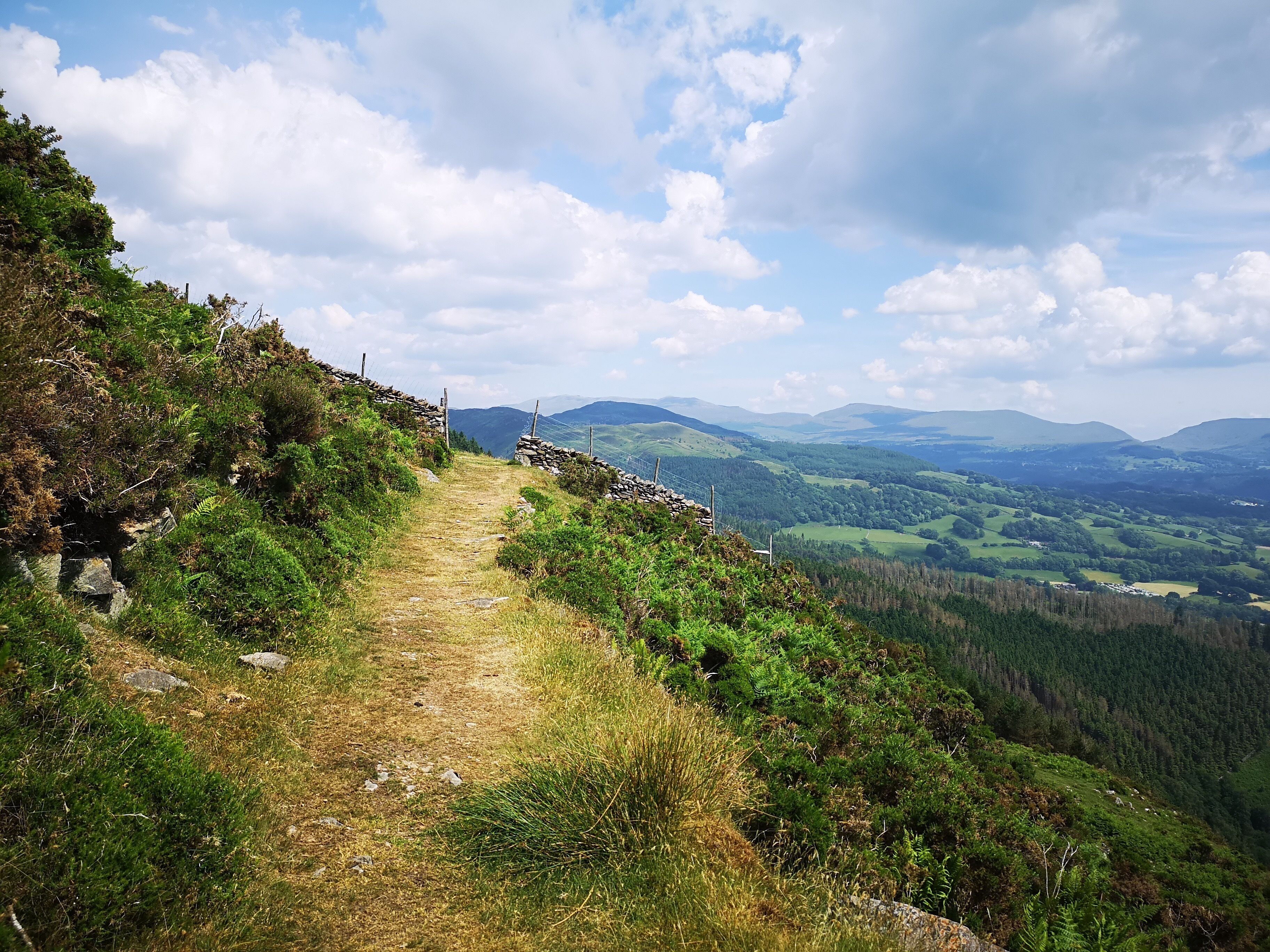 A stone wall path leading through a hilly landscape with a view of distant mountains and valleys.