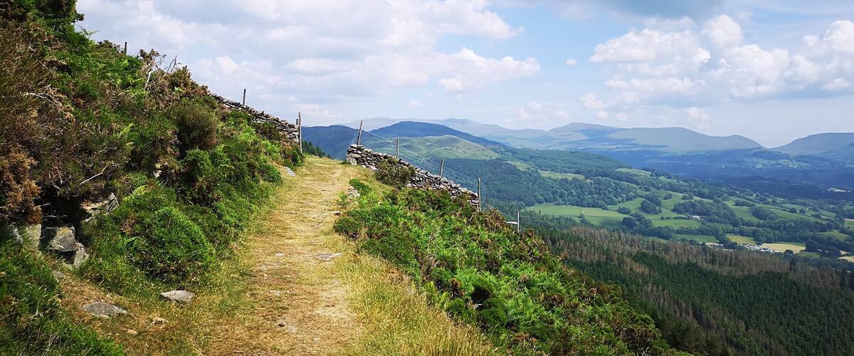 A stone wall path leading through a hilly landscape with a view of distant mountains and valleys.