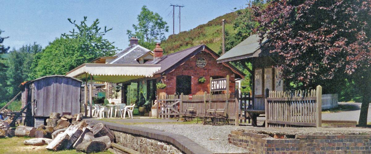 A rustic wooden building labeled "ERWOOD" with a brick facade, surrounded by a wooden fence and a small garden area.