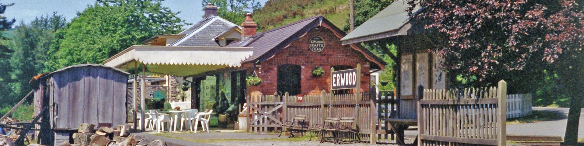 A rustic wooden building labeled "ERWOOD" with a brick facade, surrounded by a wooden fence and a small garden area.