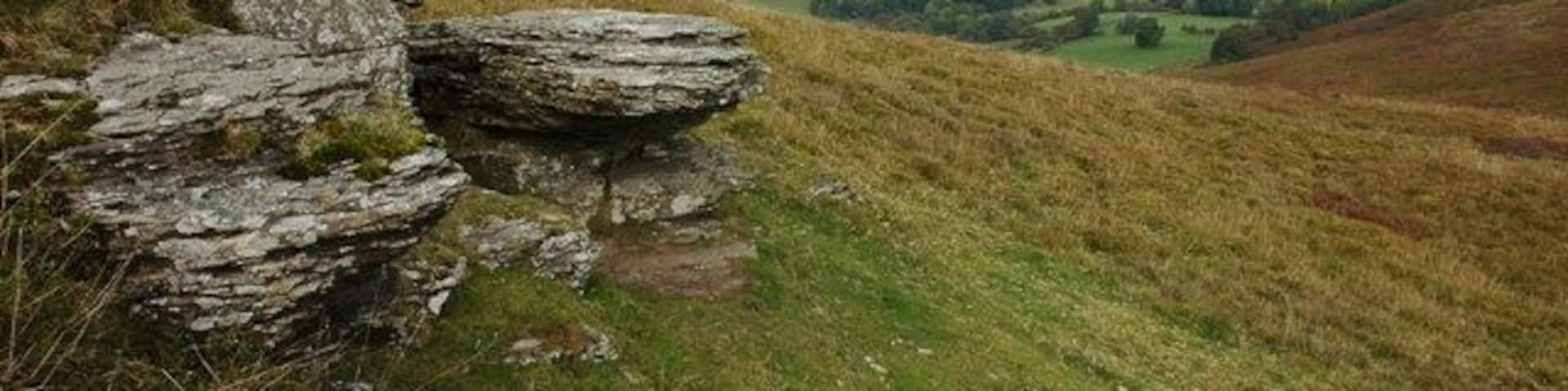 A grassy hill with large rocks and a valley with fields.